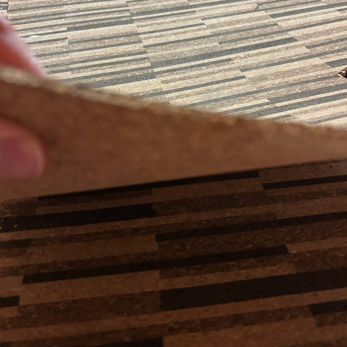 Hand lifting the corner of a Rio XL unfinished cork floor tile to show its 4 mm thickness, grain and pattern