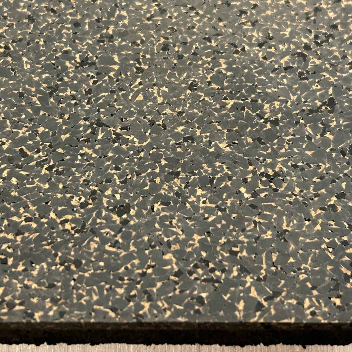 A close-up of a rubber cork tile with a  speckled surface of dark gray, black, and beige flecks, showing the dark rubber backing