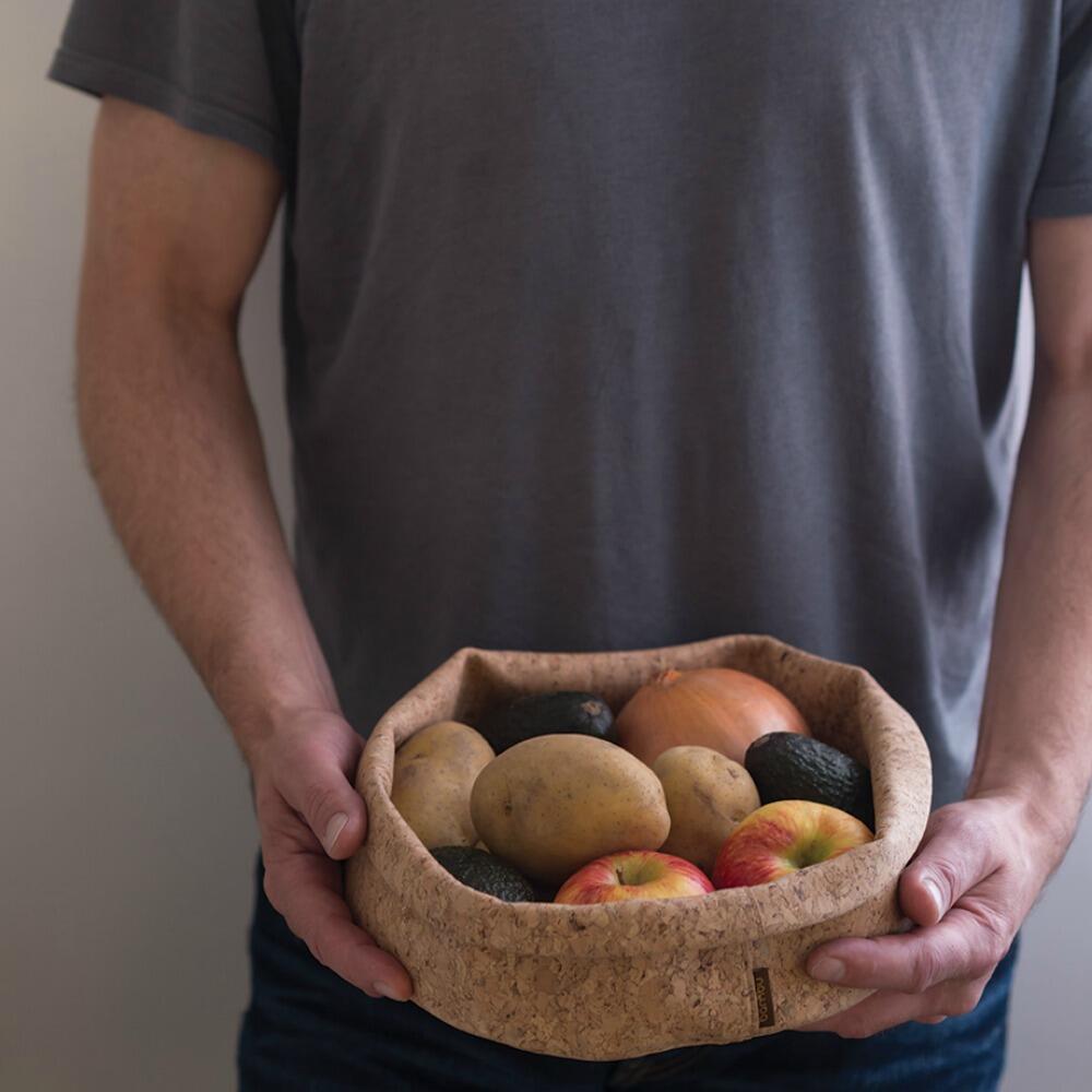 Man in a dark grey tee shirt holding a cork fabric adjust-a-bowl with folded edges holding an assortment of fruits and vegetables. 
